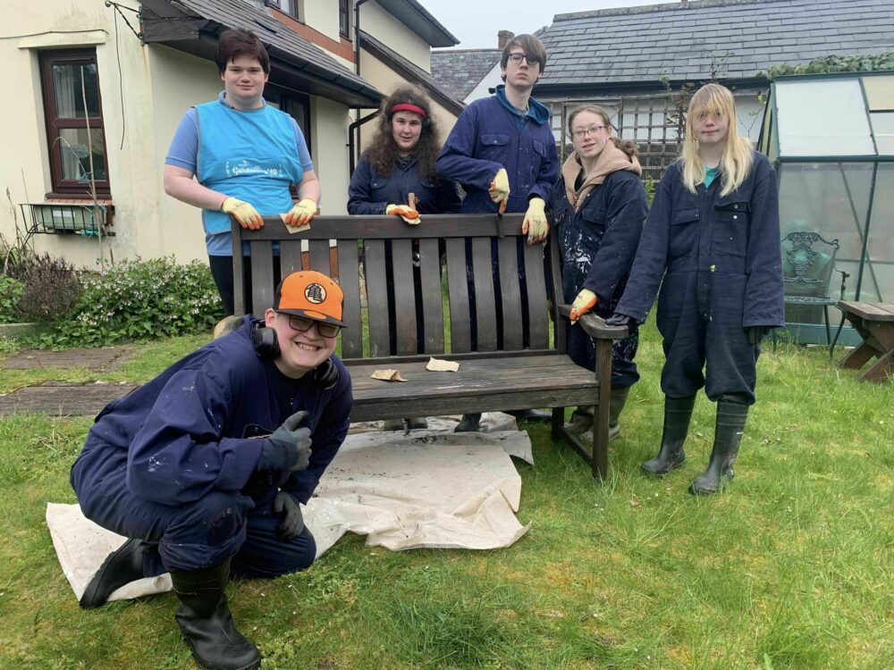 A group of young people stood around a bench in a garden, wearing work clothes