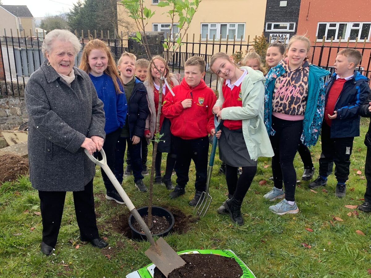 One of the committee volunteers at the Millennium Hall helps plant a tree with a group of chilldren standing beside her
